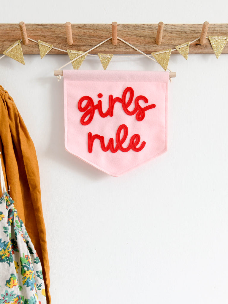 mini felt pink banner with the words girls rule sewn on in red cursive text, hung from a kids bedroom shelf.