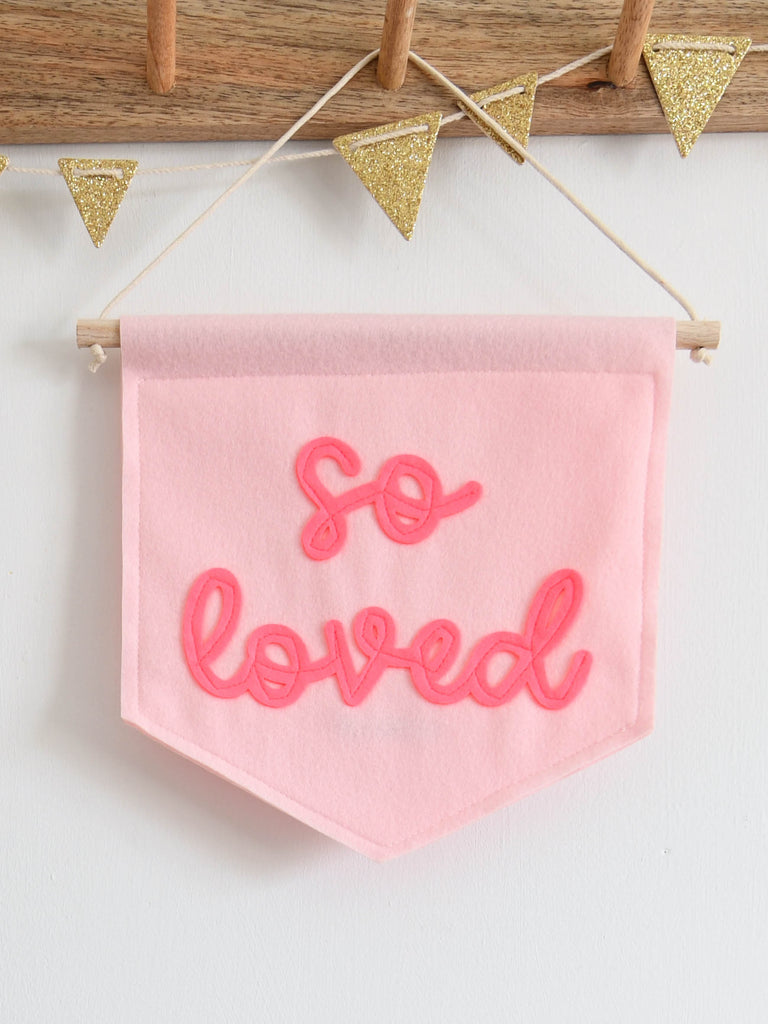pink felt banner with the words so loved sewn on in bright pink cursive text.