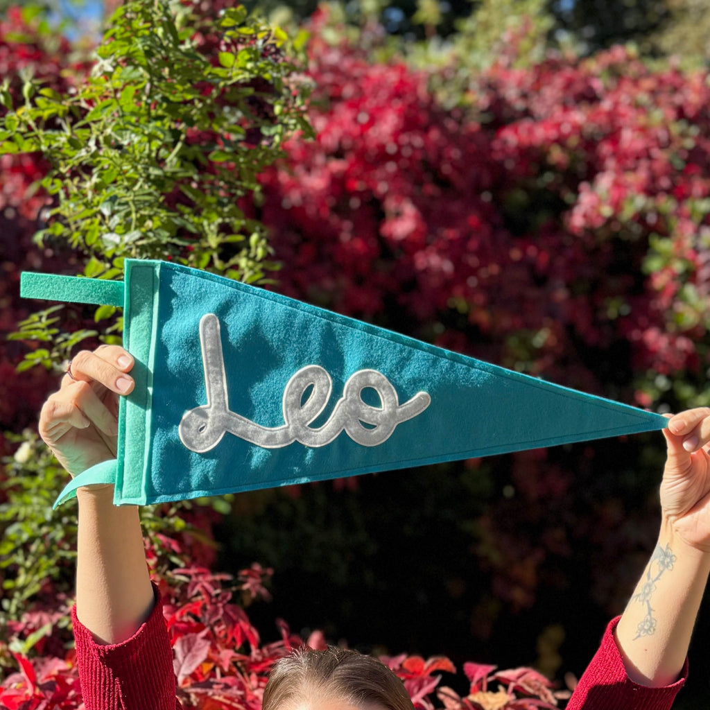 Person holding a teal pennant with 'Leo' on it against a background of green and red foliage.