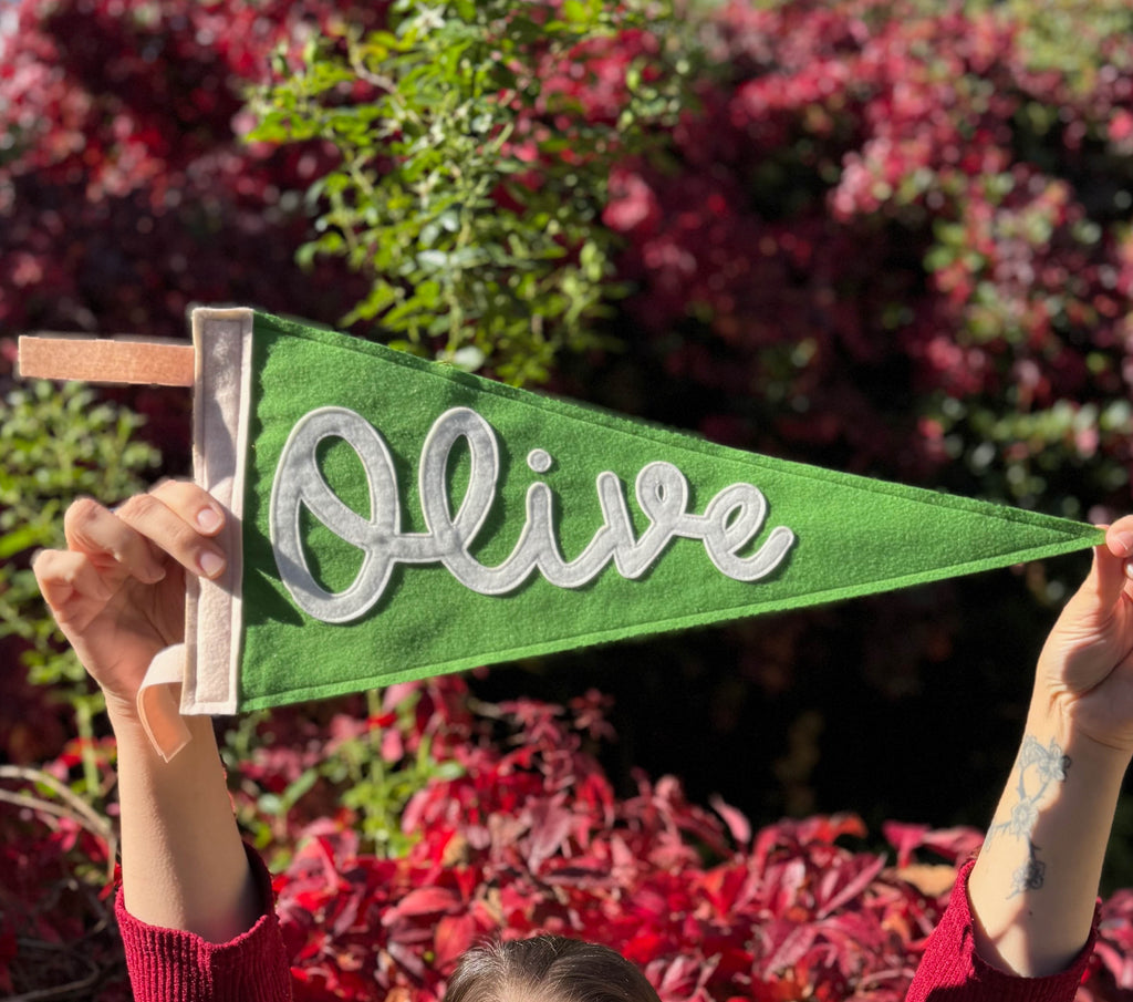 Green felt pennant with 'Olive' written on it held up against a blurred natural background