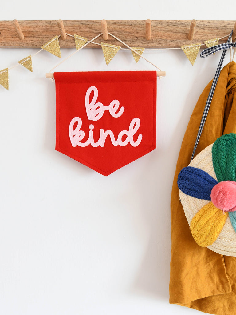a mini felt banner with the words be kind sewn on in cursive text hanging from a hook.